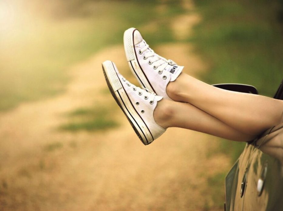 a woman wearing white sneakers hanging her legs outside of a car parked on a dirt road